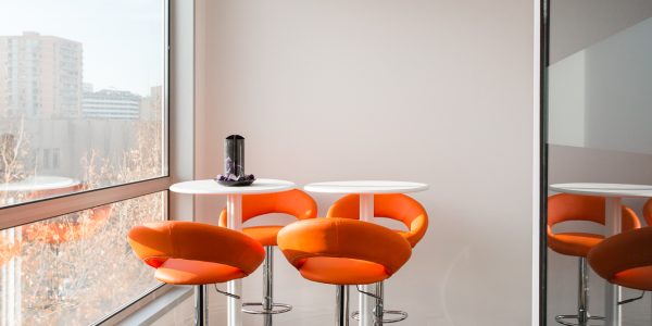 Dos mesas elevadas con sillas naranjas al lado de una ventana.
